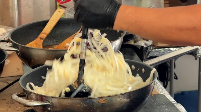 Close-up kitchen action of cook lifting mound of shredded noodles with slotted spoon above stained metal frying pan, warm busy countertop with stacked pans and vegetables