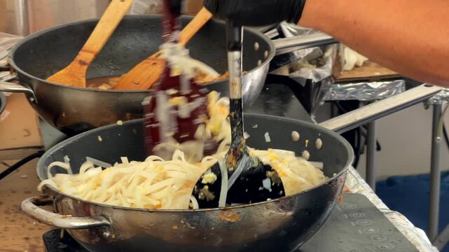 Street food noodles being lifted and transferred between metal pans with slotted spatula in gloved hands at market prep station