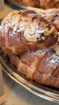 Close up almond croissant and braided brioche pastries on metal tray with spiral rolls, sliced almonds, caramelized crust, and warm bakery lighting