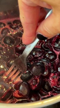 Macro close up cherries stirred and crushed in reflective metal pot with fork, releasing glossy dark purple juice and foam bubbles into bubbling deep red sauce making