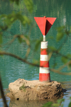 Red and white striped river navigation beacon on a stone base in the water viewed through green leaves in Phong Nha, Vietnam.