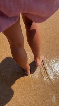 Barefoot walk along wet sandy shoreline on sunny coast, low rear view, purple skirt and carried pink shoes as surf foam sparkles nearby, leaving shallow footprints