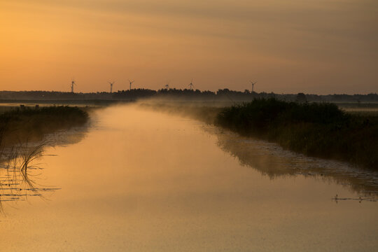 Wind turbines on the horizon over a misty river at golden sunrise. Renewable energy, green tech and sustainable landscape.