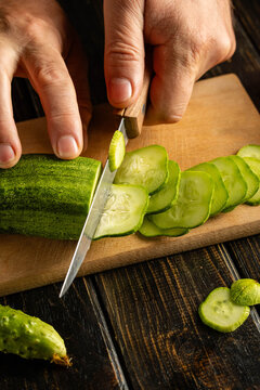 Hands slice fresh cucumber with kitchen knife on wooden cutting board. Thin green rounds rest beside blade, ready for meal preparation in close view of simple home cooking, as knife cuts through