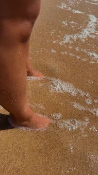 Close-up of feet and lower legs in shallow surf foam as small wave advances over sandy beach, sunlight glinting on wet skin around ankles