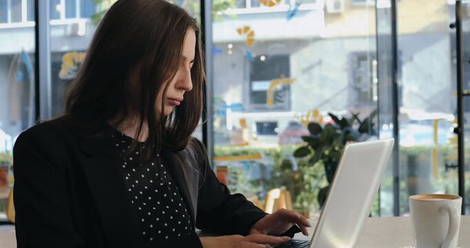 Young businesswoman works on laptop beside coffee cup inside modern cafe. Focused female professional types on portable computer while enjoying remote work routine near sunny window setting.