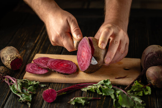 Hands slice raw beetroot with kitchen knife on wooden cutting board. Fresh beet slices reveal vivid concentric rings, creating colorful ingredients for healthy home cooking and meal preparation.