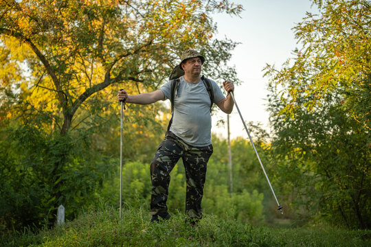 Hiker balances on grassy woodland path with trekking poles. Man in camouflage trousers and backpack wears bucket hat, holding poles wide apart with determined, steady expression under leafy trees.