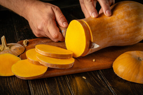 Hands are cutting butternut squash into thick slices on dark wood board. Fresh orange flesh, firm knife hold and focused food prep create warm rustic cooking moment while preparing fresh produce.