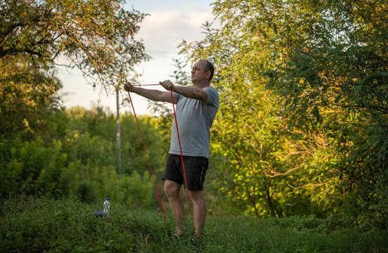Man trains with resistance band beside leafy trees, arms extended in controlled stretch. Outdoor fitness routine in summer light highlights strength work, balance, and concentration.