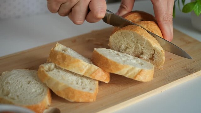 Sharp knife cuts through fresh bread loaf on wooden chopping surface. Cook methodically slices golden baguette into even pieces, demonstrating focused kitchen meal prep and culinary skill.