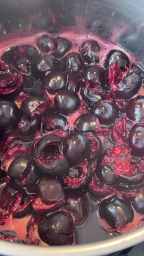 Close-up of halved dark cherries simmering in stainless steel pot, collapsing as deep red juice releases and magenta foam gathers around fruit