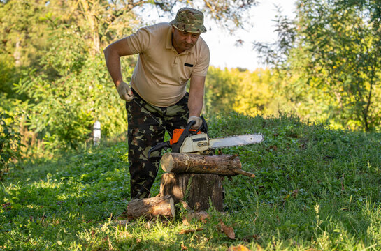 Man positions chainsaw over timber resting on tree stump in green clearing. Woodcutter in camouflage clothing and gloves looks down carefully while handling equipment for firewood preparation.