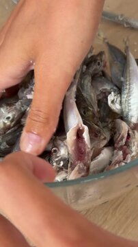Hand sorting small gutted fish in transparent bowl, pressing and sliding bodies onto wooden counter during fresh seafood preparation close overhead view with wet scales and ice crystals