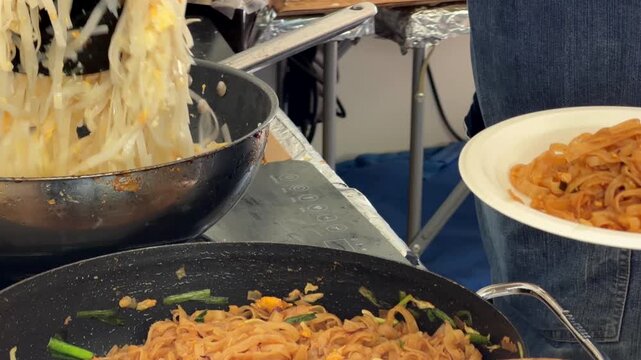 Gloved hands lifting orange-brown stirfried rice noodles from black wok to disposable plate at street food stall beside steaming bean sprouts under soft diffuse light