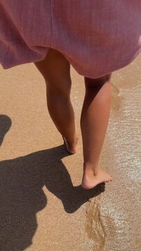 Rear close-up of barefoot legs and feet stepping on wet sandy beach near shoreline, pink garment hem visible as sunlight glints off rippled water and footprints