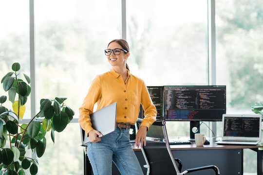 Female software developer standing in a bright modern office holding a laptop, smiling confidently beside dual monitors showing code and a large green plant