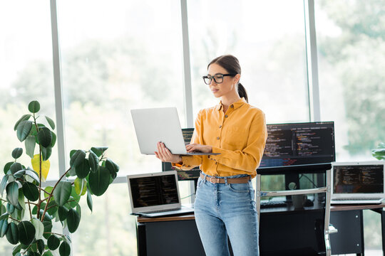 Female software developer standing in a bright modern office holding a laptop and reviewing code on multiple monitors, wearing glasses and casual yellow shirt for remote work and productivity