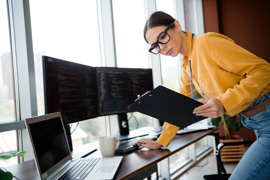 Female software developer reviewing code on a clipboard in a modern office workspace with multiple monitors, laptop and coffee while concentrating on programming and project notes
