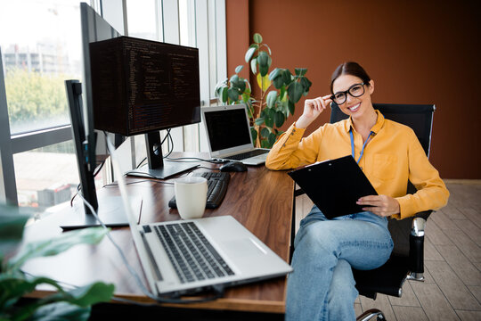 Smiling female software developer in a modern office with multiple monitors and laptop, reviewing a clipboard while working at a wooden desk