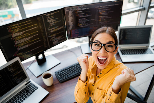 Excited female software developer celebrating coding success at her bright workstation with multiple monitors, laptops, coffee and a modern office setup