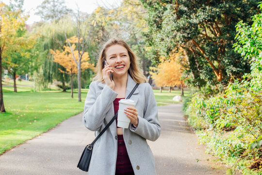 Smiling young woman has call conversation on phone while walking in autumn park and enjoying hot drink in reusable cup. Stay connected concept. Female student talking to natives while going to uni.