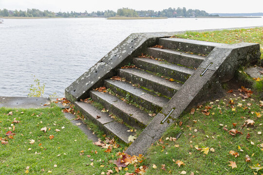 Moss-covered concrete steps descend to a quiet lakeside surrounded by green grass and scattered autumn leaves. An empty tranquil coastal scene, background photo