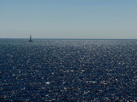 Glittering blue ocean waters clear sky and sailboat on horizon.