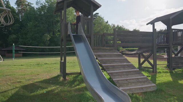 A boy sliding down a metal slide at a wooden playground on a sunny summer day