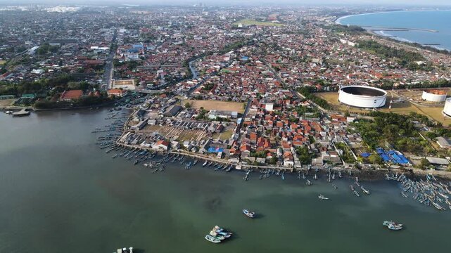 Aerial view of the coastal town of Cilacap with numerous fishing boats docked along the shore, urban rooftops, and industrial storage tanks in Cilacap Regency, Central Java, Indonesia.