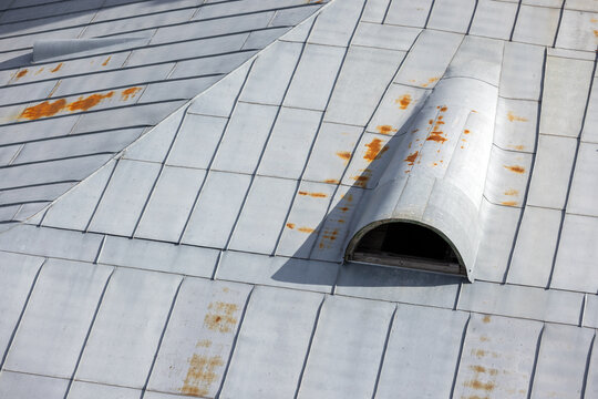 Rusted metal roof panels with a curved dormer vent reveal weathered industrial texture and corrosion details. Close-up composition emphasizes repeating lines, stains and maintenance needs