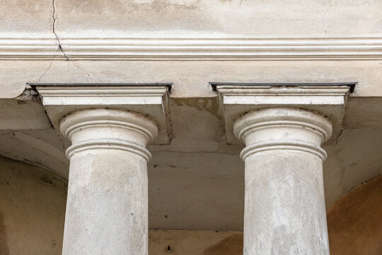 Close-up view of two weathered classical columns supporting an entablature on a historic building facade, highlighting textured stone, cracks and aged plaster that convey architectural character