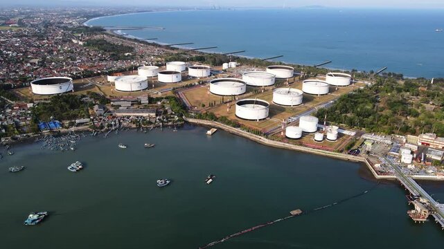 Aerial view of an industrial oil storage terminal with large white cylindrical tanks situated along the coastline, featuring small boats in the water in Cilacap Regency, Central Java, Indonesia.