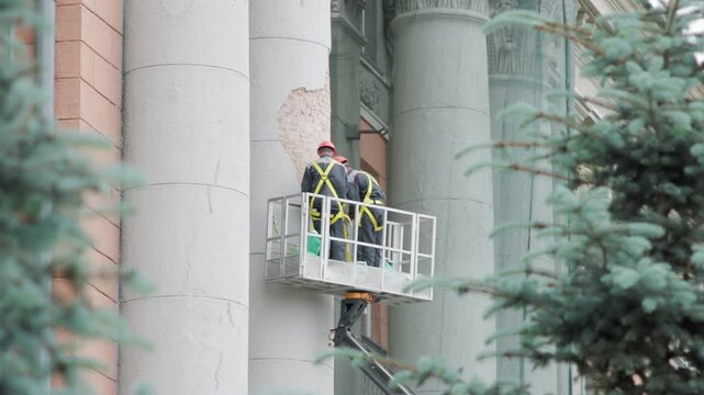 Construction workers in red helmets and yellow safety harness inspecting exposed brick and missing stucco on classical building column from aerial platform at daylight