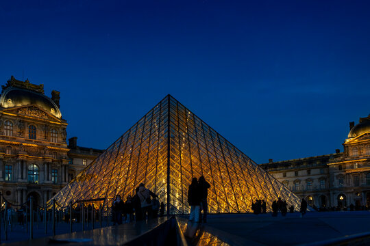Louvre pyramid with golden illumination and silhouettes of people against a deep blue sky in paris france