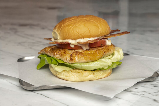 Grilled chicken sandwich with lettuce, tomato, bacon, and mayo on a marble counter with paper wrapping