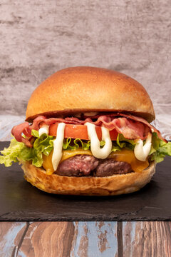 Front-facing cheeseburger with bacon, lettuce, tomato, and zigzag mayo drizzle, set on dark slate against concrete