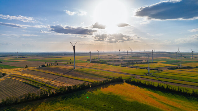 Wind turbines across farmland below clouded sunlight. Long shadows stretch across brown and yellow plots separated by rows of trees.