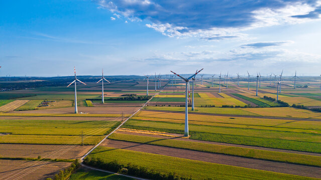 Wind turbine amid parallel crop fields. Farm tracks cross green and yellow plots around a white tower below a blue sky with thin clouds.