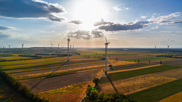 Wind turbine casting a long shadow across fields. The sun breaks through clouds above a tower beside a narrow track and rectangular farm plots.
