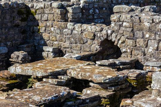 Ancient Roman stone ruin walls and raised flagstone floor foundations, showing moss and lichen on weathered masonry, outdoor sunlight