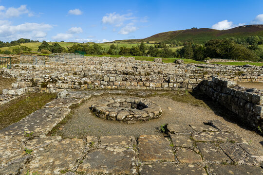 Ancient Roman stone ruins showing circular well and wall foundations, green rolling hills rising under blue sky in countryside