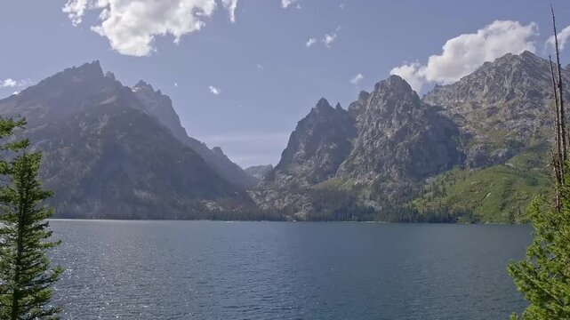 Mountain peaks rise sharply above a calm lake. Jenny Lake Finger Lake at Grand Tetons. Pines frame the view, gently swaying in the breeze. Sky is bright, with soft clouds drifting lazily