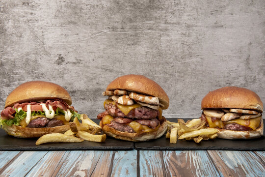 Three different gourmet burgers lined up on slate boards with fries against a weathered concrete backdrop