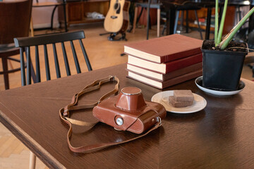 Vintage camera in leather case with strap on wooden table beside books and biscuits. Analog photography equipment. Creative studio workspace. Reading break and music room interior. © Анастасия Бурлакова