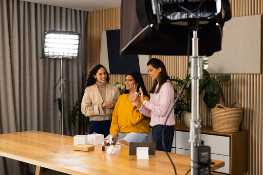 Diverse female creators testing skincare on wooden table in studio, mustard top model reacting