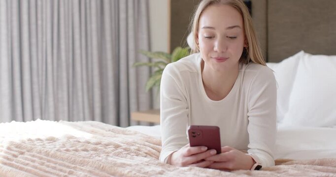 Woman in early twenties reclining on bed, holding red smartphone, scrolling and typing messages