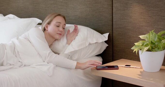 Adult female lying in bed reaching smartphone on bedside table as alarm ringing, tapping to snooze