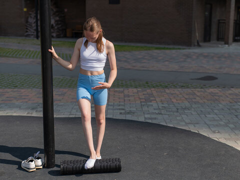 A Caucasian woman stretches her feet with a foam roller at a sports ground.
