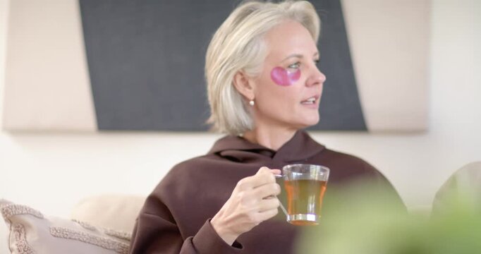 Senior woman hearing offscreen voice, turning toward it with glass mug on sofa, brown pullover
